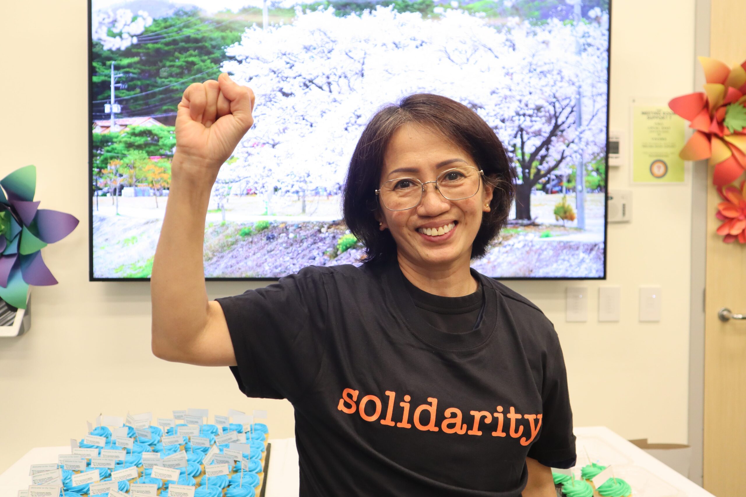 A woman with a raised fist wearing a t-shirt that says “solidarity.” She is standing in front of a television and a table. The table has blue and green cupcakes on them.